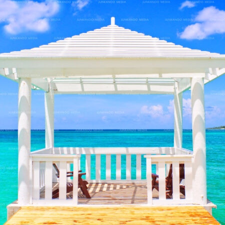 A Gazebo in the middle of the ocean in The Bahamas beneath while puffy cumulous clouds and above green and turquoise water.