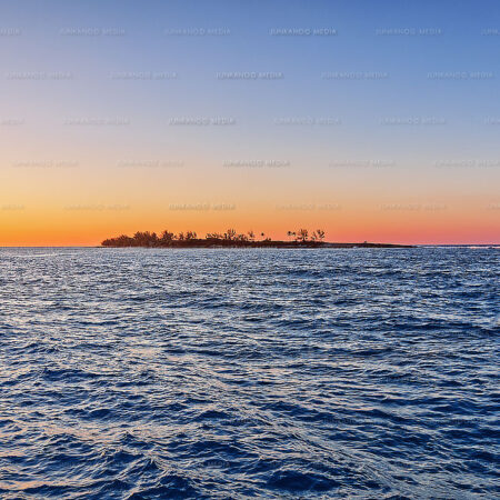 An island sits in the soft glow of sunset surrounded by ocean waves.