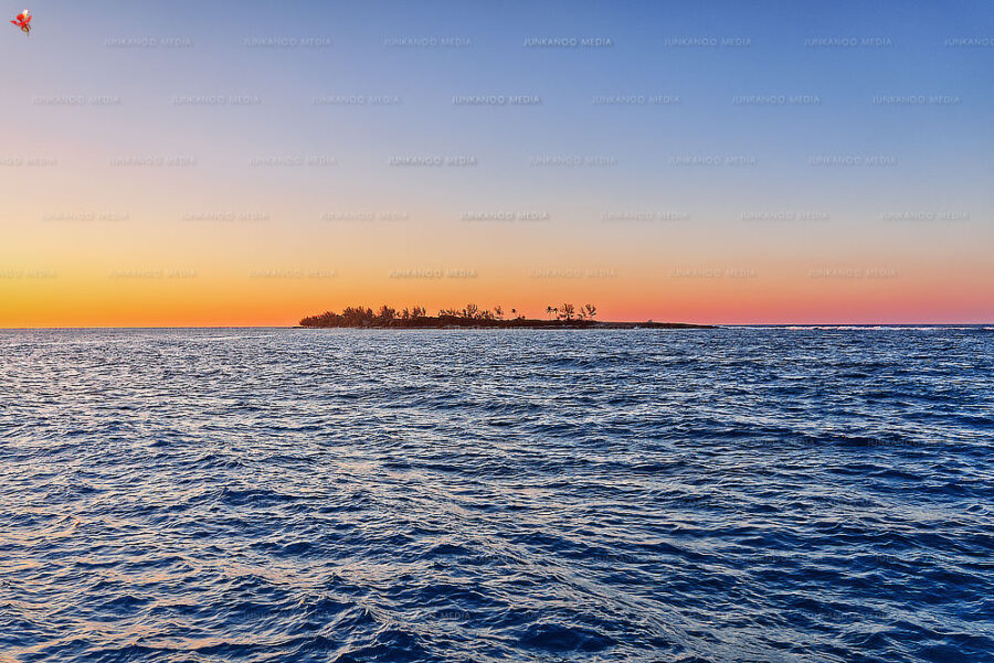 An island sits in the soft glow of sunset surrounded by ocean waves.