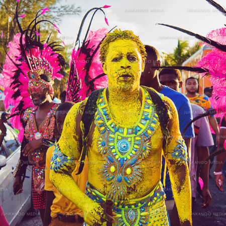 A man in full-body yellow paint walks along Saunders Beach in Nassau Bahamas in front of people in Junkanoo Costumes.