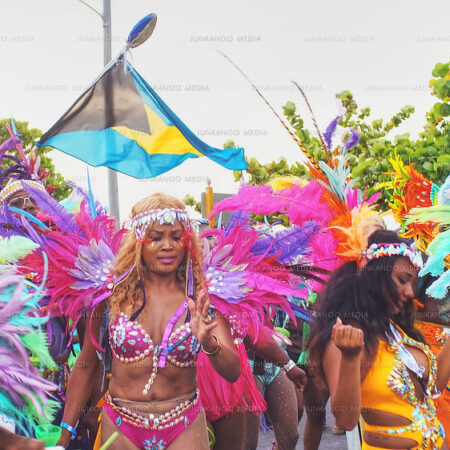 Three women in Junkanoo Costume dance on West Bay Street in front of the Bahamian flag, surrounded by flamboyant colors.