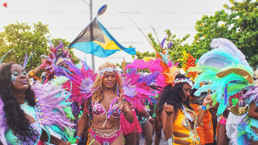 Three women in Junkanoo Costume dance on West Bay Street in front of the Bahamian flag, surrounded by flamboyant colors.