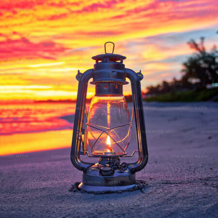 A lantern sits on the sand beneath pink, orange and yellow clouds lit by the sunrise.