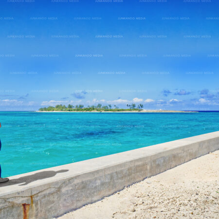 A man stands at the extreme western end of Arawak Cay looking toward the near horizon where an island in The Bahamas is situated.